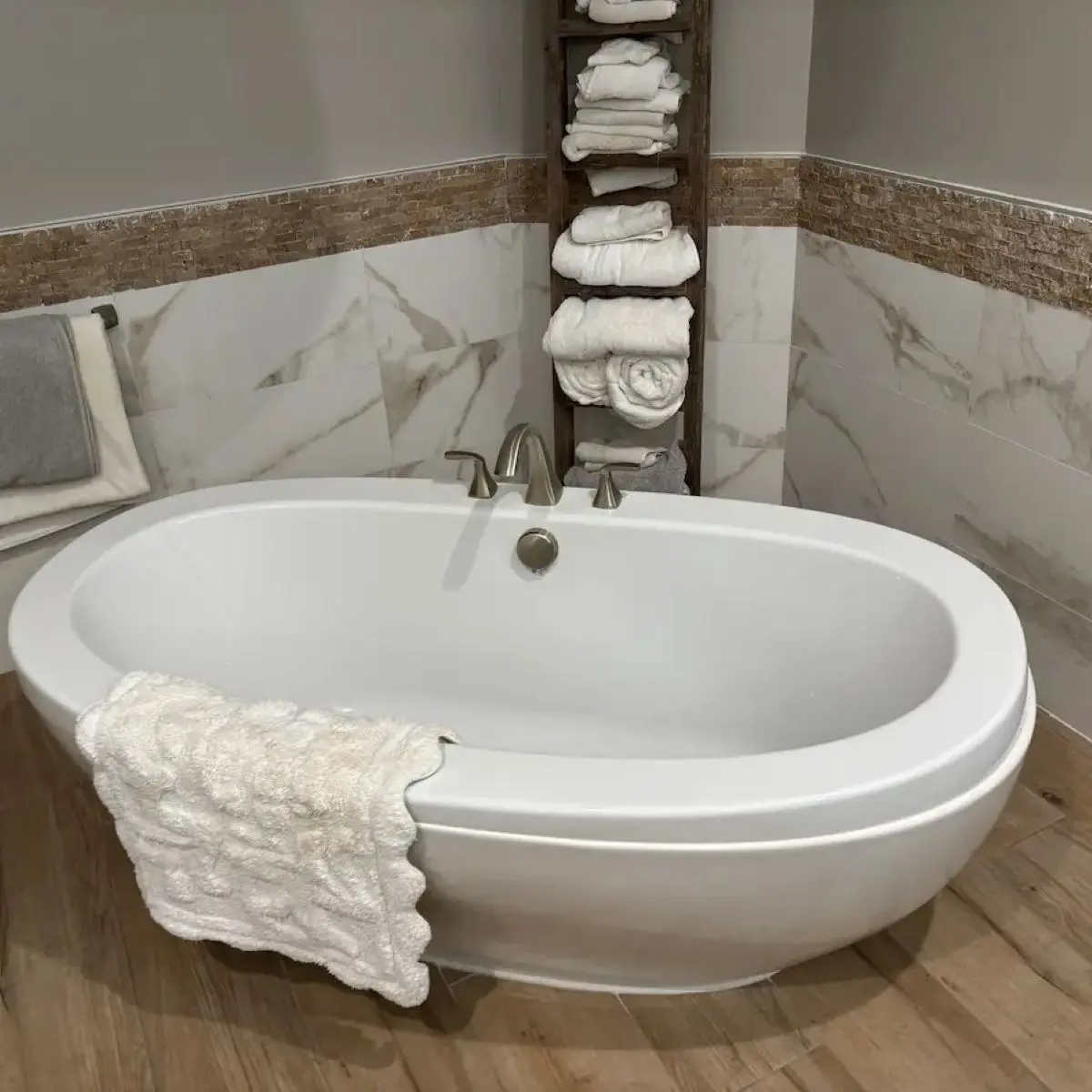 A large white oval freestanding bathtub with a brushed nickel faucet set against marble-look wall tiling and wood flooring.