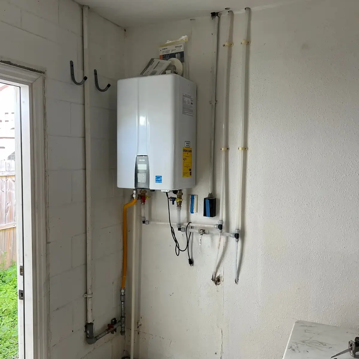 A wall-mounted white Navien tankless water heater inside a utility space with a yellow flexible gas line and clean water inlet and outlet pipe runs.